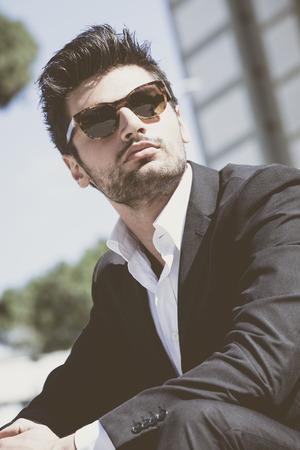 Confident young successful man sitting in the city. Wearing sunglasses, behind him at modern office building. Trendy hair, white shirt and jacket.の写真素材