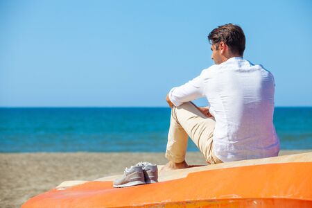 Lonely man on the beach above a boat looking at the sea. Seated shoulders on the orange boat. The young man wears a white shirt and a brown trousers. His shoes are resting on the sling.の写真素材