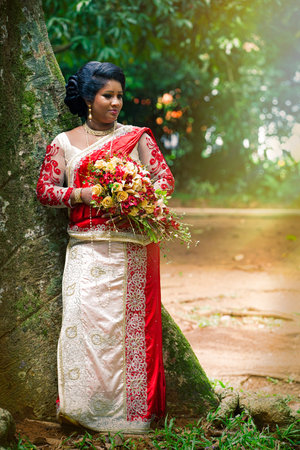 Kandy, Sri Lanka. July 24, 2016: An indian bride in a park near a tree in Kandy in Sri Lanka. She wears Saree typical Indian bridal dress women. With bouquet of flowers in hands.のeditorial素材