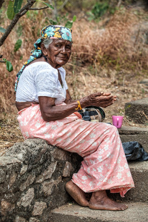 Dambulla, Sri Lanka. July 22, 2016: An Elderly woman in Dambulla in Sri Lanka with hands asking for charity is sitting on a low wall on the street. She wears clothes of Asian culture.のeditorial素材