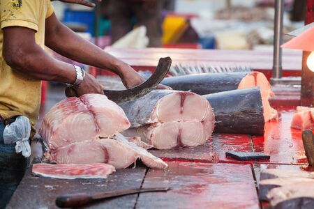 Hands with knife chopping fish outdoors. A fisherman and fish seller is tracking sliced a big fish on a wooden table. Blood shed on the wooden table. Fish market in Negombo, Sri Lanka.の写真素材