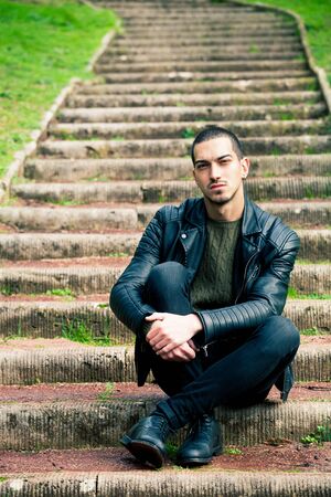 Handsome young boy sitting on a staircase in a natural park. Crouch, rock style clothes with leather jacket and dark jeans. Short hair and earring.の写真素材