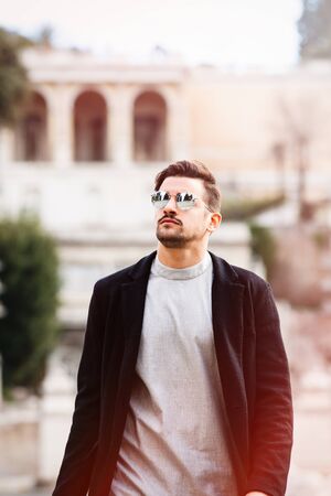 Stylish man walking with sunglasses and modern hairstyle and beard, looking up. Outdoor in the street. White t-shirt and black jacket.の写真素材