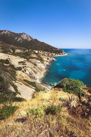 Seashore coastline with beach and rocks and rocky slope of the Island of Elba in Italy. Many people on the beach sunbathing. Blue sea with aerial view. Dwellings of a small village.の写真素材
