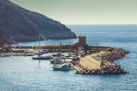 Elba Island, Italy. June 25, 2016: Small marina in the town of Marciana Marina on the Island of Elba in Italy. Some boats moored in the sea under a rocky wall of a mountain in the background. Rocks and slopes on the sea.のeditorial素材