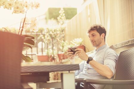 Outdoor man sitting and relaxed texting with a smartphone in his hands. Illuminated environment, man wears a heavenly shirt. Spring scene. Social connection.の写真素材