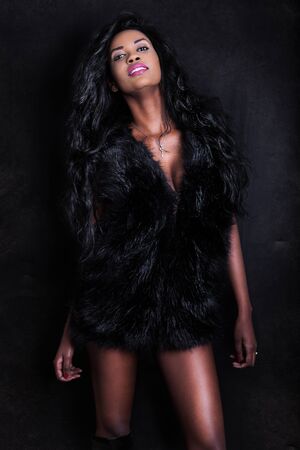 Beautiful young African American seductive black model woman on dark background. An attractive young woman wearing a furry black vest. Sensual look and perfect physique.の写真素材