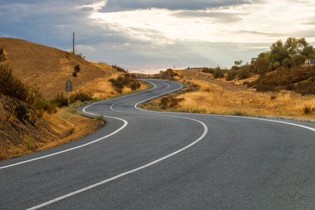 Asphalt road in the mountains at sunset. Spain, Andalusia. Secondary paved road with curves. Rural road on a cloudy summer day. El Granado, Spain. HU-6400 country road.の写真素材