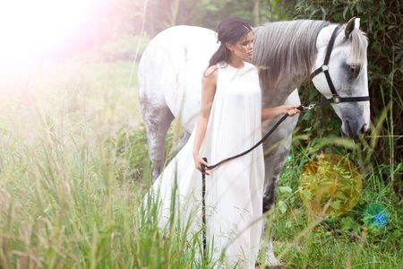 An attractive young woman in a white, flowing dress guides a white horse through an idyllic meadow. There is a lighting effect in the upper left corner of the image. Horizontal shot.の写真素材