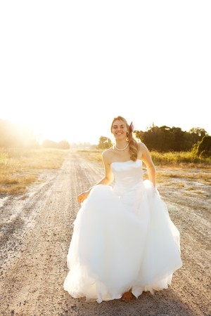 An attractive young bride wearing a white wedding dress is standing in the middle of a dirt road in a rural landscape. Vertical shot.の写真素材