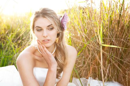 An attractive young bride wearing a white wedding dress and pearls is sitting with her head on her hand in the grass. Horizontal shot.の写真素材