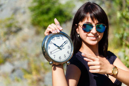 Beautiful girl in a black top and sunglasses showing a clock. Focus on a clock, concept of time to...