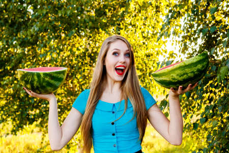 Beautiful blonde girl in a blue dress with long hair holding two halves of a sliced watermelonの写真素材