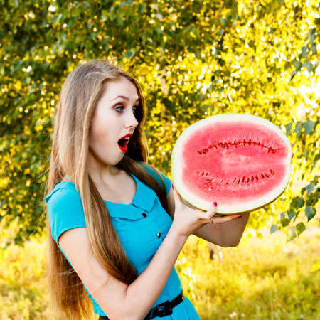 Beautiful blonde girl in a blue dress with long hair holds a half of watermelon. backlight, outdoors.の写真素材