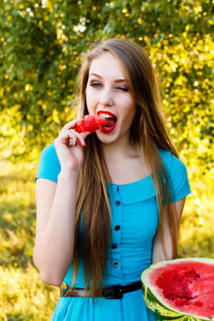 Beautiful blonde girl in a blue dress with long hair eating a watermelon outdoorsの写真素材