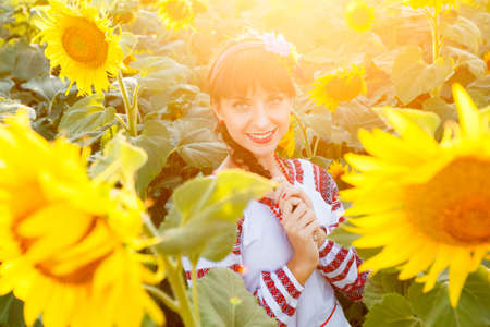 Pretty young woman in national ukrainian blouse embrodery looking to camera on a sunflower plant at...