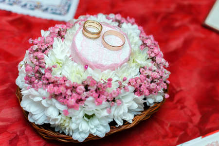 Two gold wedding rings on Beautiful white and pink bouquet in rustic basketry. Red fabric background. Close up.の写真素材