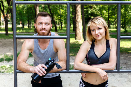 Sporty blonde girl and bearded man is resting after workout training in a park outdoor. Man holding a black bottle with water. Concept of a healthy lifestyle, losing weight.の写真素材