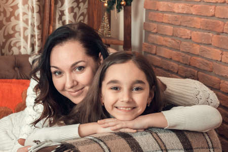 Two beautiful girls, mother and daughter siting on a sofa in Christmas decorated room. Looking at camera.の写真素材