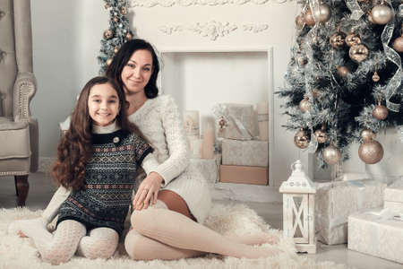 Two pretty girls, mother and daughter siting on a floor in Christmas decorated room. Family celebrate xmas eve. Smiling, Looking at camera.の写真素材
