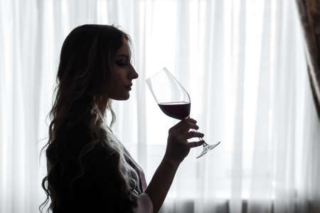 Sensual young woman dressed in a robe drinks a  red wine from big wineglass, silhouette in a backlight on a window backgroundの写真素材