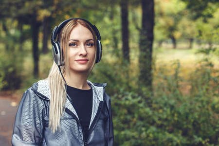 beautiful young blonde woman walking on city park, enjoys listening to music on headphones. Looking at camera.の写真素材