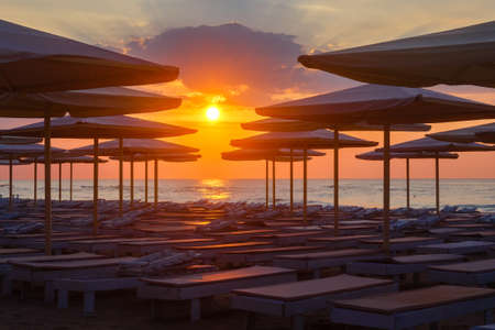 Silhuettes of beach loungers and umbrellas on a deserted beach in the evening on a sunset backgroundの写真素材