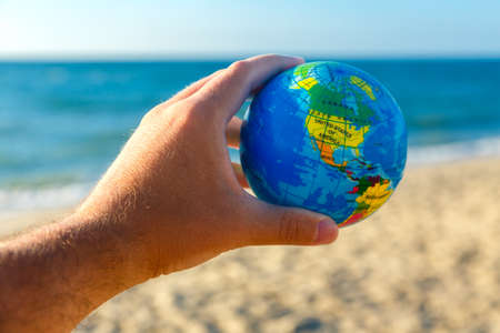 Male hand holds globe of planet Earth on a seaside background. Travel concept.の写真素材