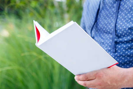 Man student dressed in a blue shirt holds open white empty book outdoors. Hands closeup on a...