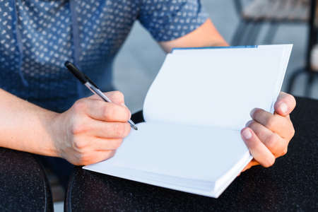 Man sitting at the table in a cafe, writing on the white empty pages of opened  book closeup.
