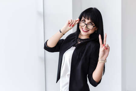 Portrait of young beautiful smiling woman with glasses standing near the window, looking at camera, showing thumbs up, sign victory.の写真素材
