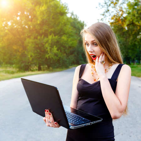 Beautiful blonde female manager with open mouth in surprise looks to laptop, working in a park outdoors on a sunset.の写真素材