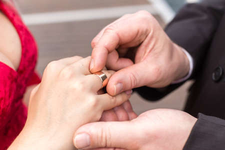 Groom puts a wide wedding ring on the finger of Bride, dressed in a sexy red dress with a decollete.の写真素材