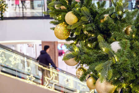 Golden Christmas balls and garland on a decorated fir tree in Shopping Mall. Blurred people on escalator, in festive business center make New Year's purchases,.の写真素材