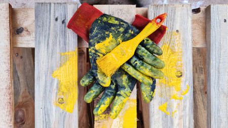 Black work protective gloves and a paintbrush smeared with yellow paint lie on a wooden pallet.の写真素材
