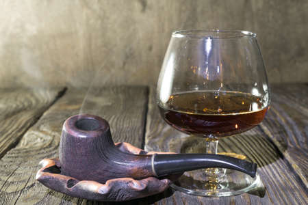 A Smoking pipe in the ashtray and a glass of cognac on oak textured tableの写真素材
