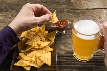 Wooden table male hands holding a mug with beer and nachos dipped in the sauce.の写真素材