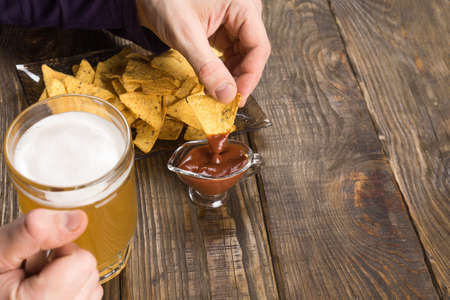 Wooden table male hands holding a mug with beer and nachos dipped in the sauce.の写真素材