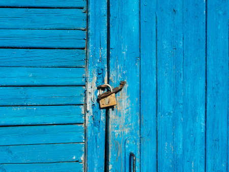 The wooden door with peeling blue paint is locked with a padlockの写真素材