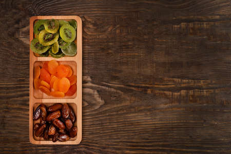 Dates, dried apricots and kiwis in a Compartmental dish on a dark wooden tableの写真素材