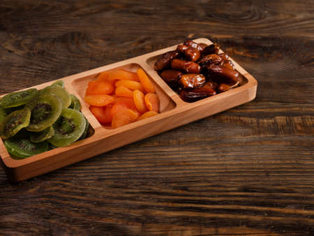 Dates, dried apricots and kiwis in a Compartmental dish on a dark wooden tableの写真素材