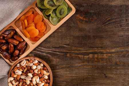 Dates, dried apricots and kiwis in a Compartmental dish and assortment of nuts in wooden bowl on a dark wooden tableの写真素材