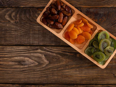Dates, dried apricots and kiwis in a Compartmental dish on a dark wooden tableの写真素材