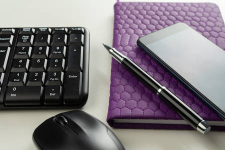 White wooden table with keyboard, pen, mouse, notebook, mobile phone.
