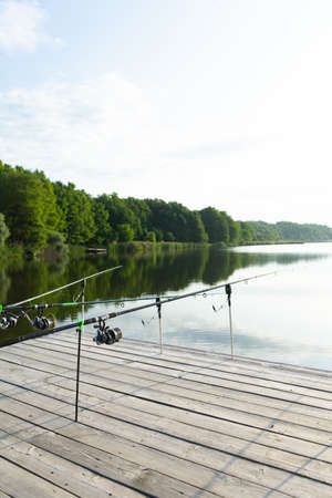 Carp fishing on beautiful blue lake with carp rods and rod pods in the summer morning. Fishing from the wooden platformの写真素材