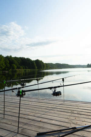 Carp fishing on beautiful blue lake with carp rods and rod pods in the summer morning. Fishing from the wooden platformの写真素材