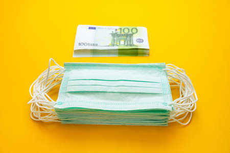 A stack of medical masks next to a stack of money on a yellow background. The concept of increasing the price of surgical masks due to the rapid spread of the coronavirus.の写真素材