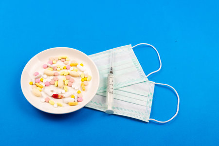 A variety of pills on a plate, medical masks and syringes next to each other on a blue background...