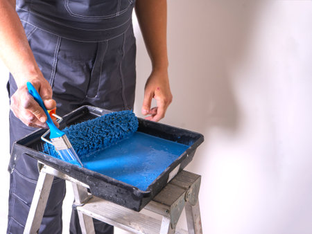 The painter dips his brush in paint bath of blue paint. Cuvette for rollers on a stepladder.の写真素材