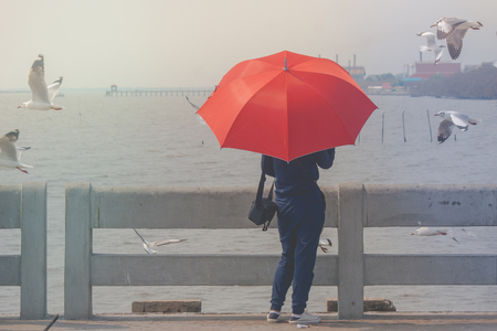 Woman standing on concrete bridge, she holding red umbrella and looking at sea and seagulls flying at Bangpu Recreation Center, Smutprakarn Province, Thailand.の写真素材